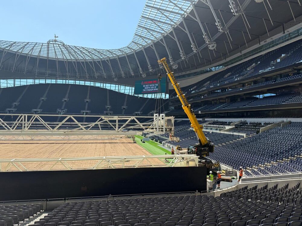 A compact city crane working in the bowl of Tottenham Hotspur Stadium, tiered seating sweeping behind