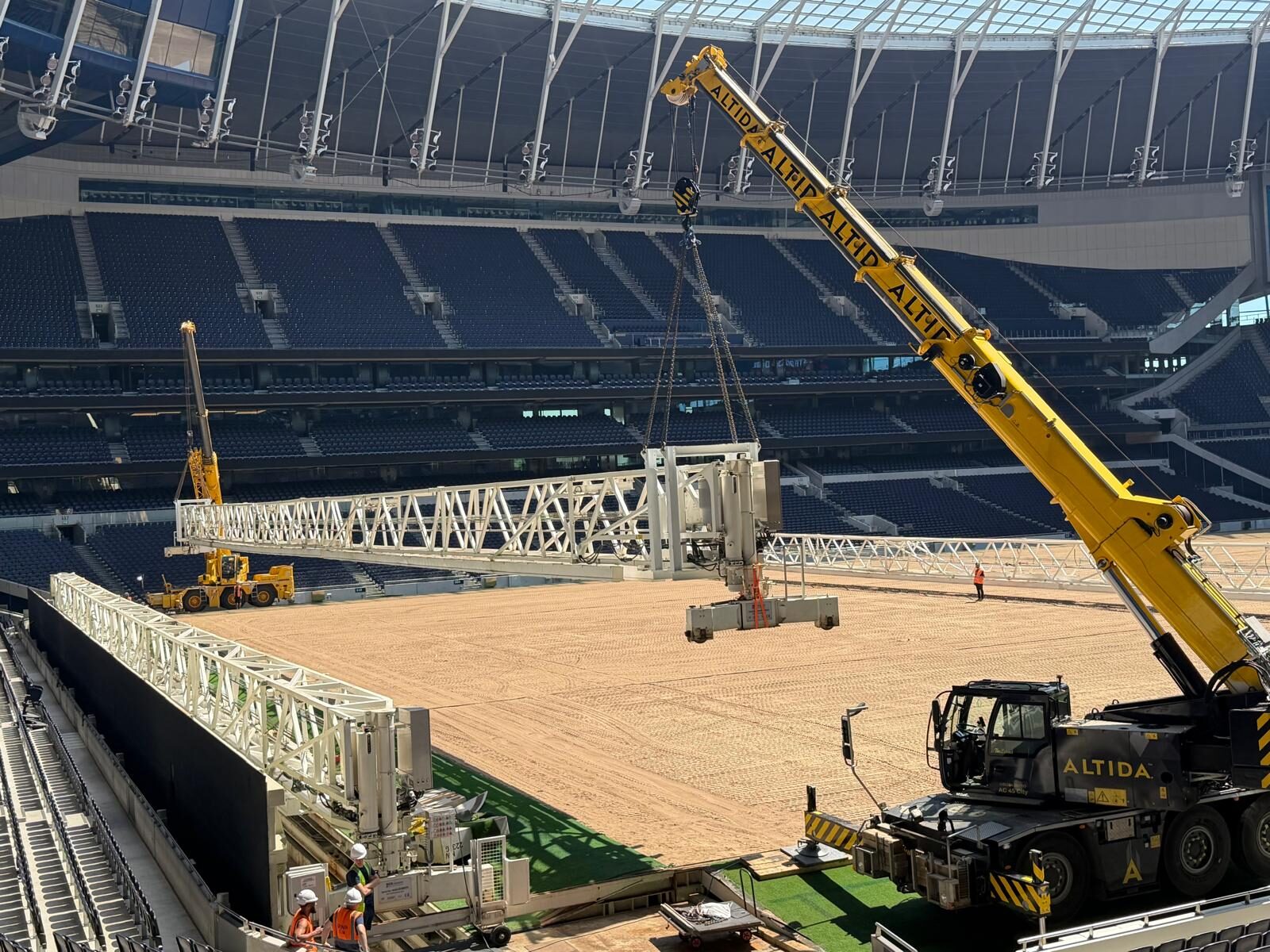 Two Altida cranes lifting a 60-metre grow-light bracket in tandem over the pitch at Tottenham Hotspur Stadium